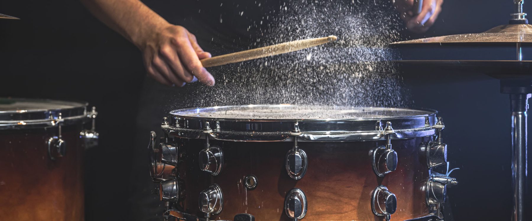 A male drummer plays snare drum with drumsticks in a dark room.